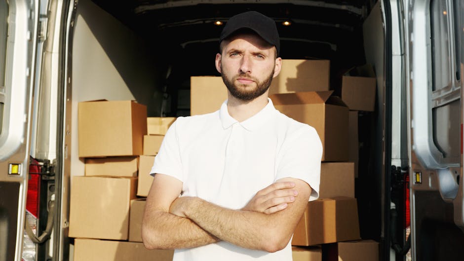 A man standing beside a van during a home relocation process, wearing a grey knit beanie, a green padded vest over a navy blue jacket with white stripes along the sleeves, and holding a black strap or