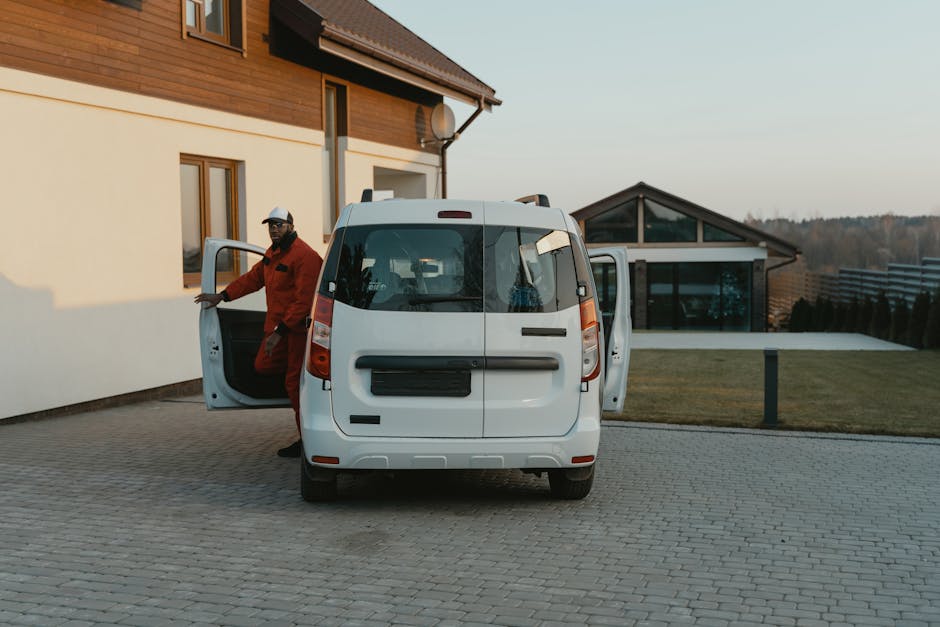A man wearing a red jacket, black trousers, and a white helmet is standing outside a white panel van with its side door open, preparing for a home relocation. The van is parked on a paved driveway adjacent to a modern residential building with cream-colored walls and wooden cladding, and it has a large rear window and black bumper. Inside the open side door of the van, packing materials and cardboard boxes are visible, indicating the loading process involved in furniture transport and moving logistics. The man appears to be either loading or unloading items, with the background showing a contemporary glass-fronted house and a landscaped garden, under natural daylight. Man and Van Charlton's services relate to packing and moving, emphasizing efficient furniture transport and house removals in the Charlton station area SE7.