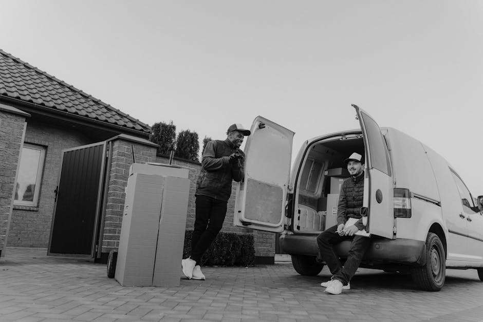 Two men are engaged in home relocation activities outside a residential property as part of furniture transport and packing and moving procedures. One man, standing beside a large cardboard box and a furniture trolley, is wearing a cap, dark jacket, and white sneakers, and appears to be inspecting or communicating. The other man, sitting on the open rear door of a white cargo van with its interior visible, is dressed in a hoodie, jacket, and hat, and is smiling at the camera. The van, parked on a paved driveway, is in the process of being loaded or unloaded, with cardboard boxes and packing materials possibly inside. The background includes a brick house with a tiled roof and a small window, along with some trees and bushes. The setting is outdoor during daylight, with natural light illuminating the scene, which embodies typical elements of house removals and relocation services carried out by [COMPANY_NAME] in the Charlton area.