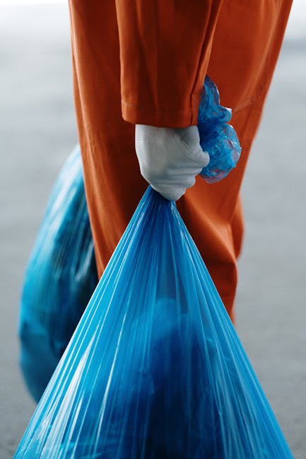A worker in a yellow and red high-visibility jacket is standing behind a large red industrial waste collection vehicle positioned on the side of a street during daylight hours. The rear of the vehicle