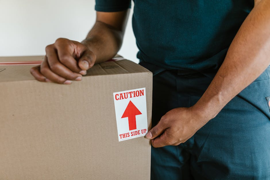 A man with dark, curly hair and a beard stands indoors against a plain white background, holding a cardboard box labeled 'OFFICE' with both hands. He is wearing a red long-sleeved shirt and greenish-g
