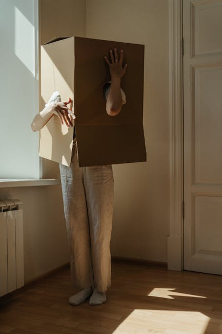 A person standing indoors near a window with sunlight casting shadows on the wooden floor, wearing beige trousers and white socks. They are holding a cardboard box with their left hand, while their right arm is inserted into an opening cut into the front of the box, resembling a makeshift costume or hiding position. The box appears to be plain brown corrugated cardboard, with the person's hand visible through the hole. The setting features a white radiator below the window and white-paneled doors adjacent to the individual. This scene illustrates the packing or unpacking process involved in home relocation, which is a typical service offered by companies like Man and Van Charlton. The environment suggests a residential property prepared for a furniture transport or loading process, with natural light and minimal clutter, aligning with house removals and moving logistics.