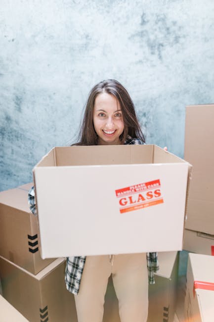 A woman with long brown hair smiling while holding an empty cardboard box labeled 'Glass' in a modern interior space with a blue textured wall. She is dressed in casual clothing and standing among other packed boxes made of cardboard, some with black and red tape, suggesting an ongoing home relocation or packing process. Behind her, additional boxes are stacked or placed nearby, indicating preparatory activities for furniture transport or moving logistics. The scene is well-lit, highlighting the careful packing materials such as cardboard and tape used during furniture transport, characteristic of professional removals services like those provided by Man and Van Charlton. This image visually captures the packing and loading process involved in house removals, supporting tips to avoid hidden charges during a move.