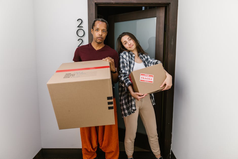 A person standing indoors near a window with sunlight casting shadows on the wooden floor, wearing beige trousers and white socks. They are holding a cardboard box with their left hand, while their right arm is inserted into an opening cut into the front of the box, resembling a makeshift costume or hiding position. The box appears to be plain brown corrugated cardboard, with the person's hand visible through the hole. The setting features a white radiator below the window and white-paneled doors adjacent to the individual. This scene illustrates the packing or unpacking process involved in home relocation, which is a typical service offered by companies like Man and Van Charlton. The environment suggests a residential property prepared for a furniture transport or loading process, with natural light and minimal clutter, aligning with house removals and moving logistics.