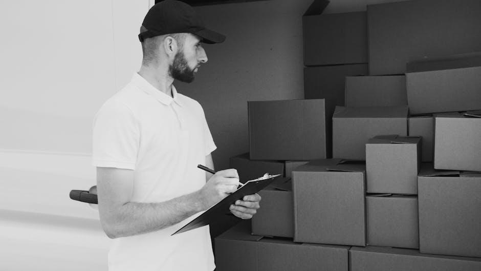 A man in a white polo shirt and black cap is standing inside a property, writing on a clipboard as part of a house removal process. Behind him, there are numerous medium-sized cardboard boxes stacked
