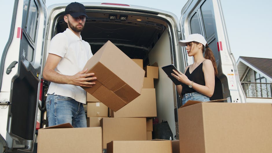 A man wearing a black cap, white T-shirt, and white wireless earbuds is inside the open back of a large moving van, which is parked on a pavement and filled with numerous stacked cardboard boxes of va