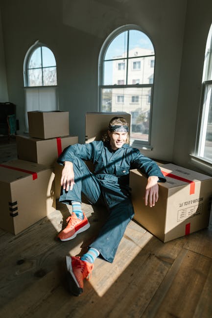 A person wearing a white T-shirt and blue jeans is kneeling indoors on a concrete floor, using a red marker to write on a cardboard box labeled 'Items.' Several other cardboard boxes, some open and so